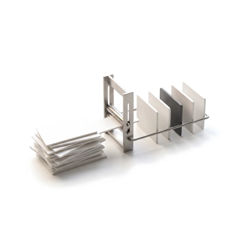A loose stack of ceramic sheets fed through a steel press, coming out as a neat row of upright cards, one graphite.
